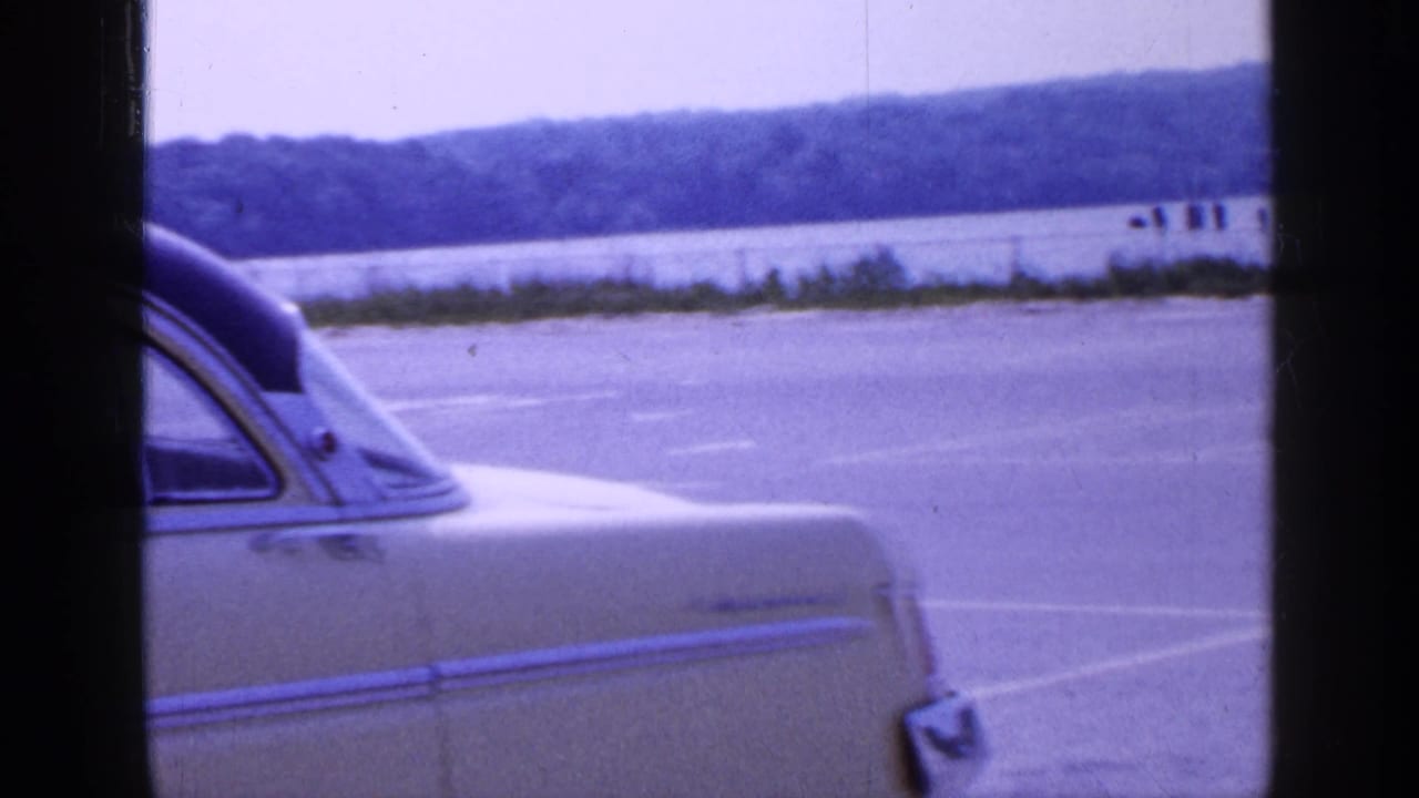 1965 Long Island frame showing the rear portion of the light-colored sedan near water and a distant tree-lined shore.
