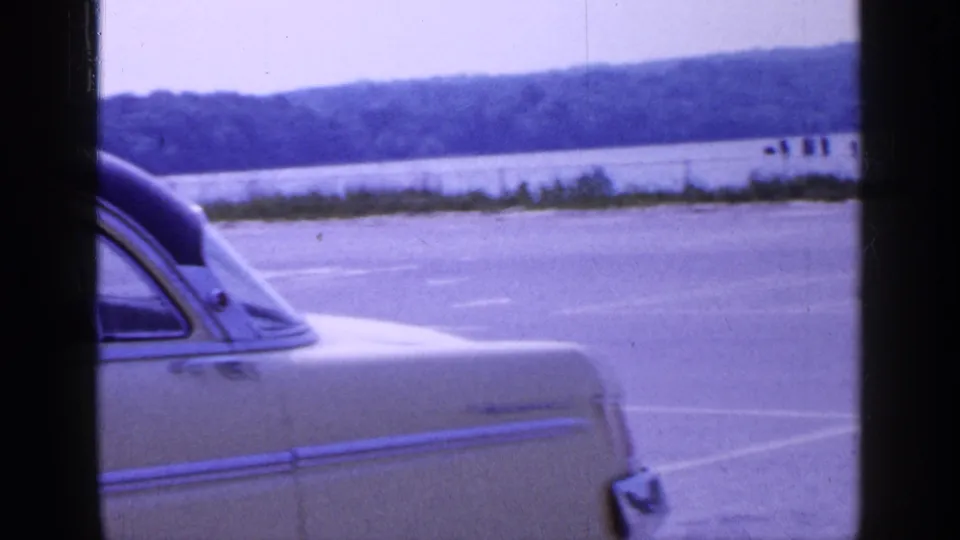1965 Long Island frame showing the rear portion of the light-colored sedan near water and a distant tree-lined shore.