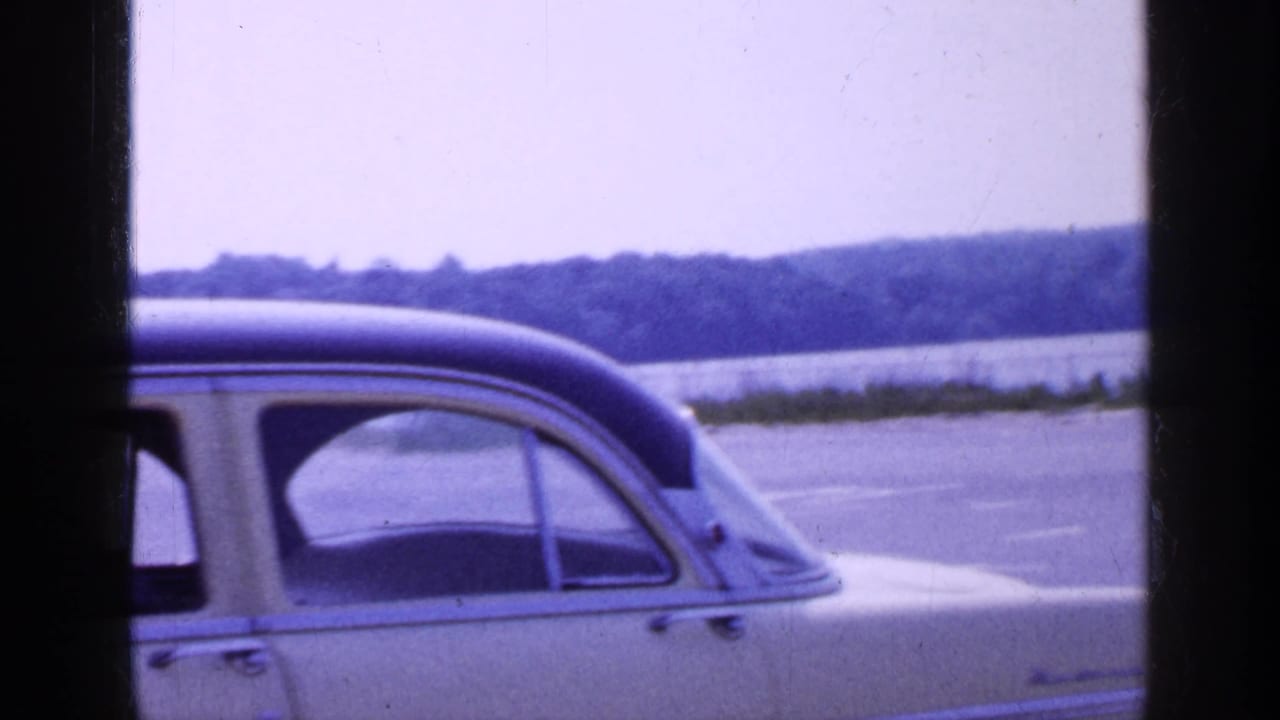 1965 Long Island home-movie frame showing a cream four-door sedan by a waterfront, partly obscured by film gate edges.