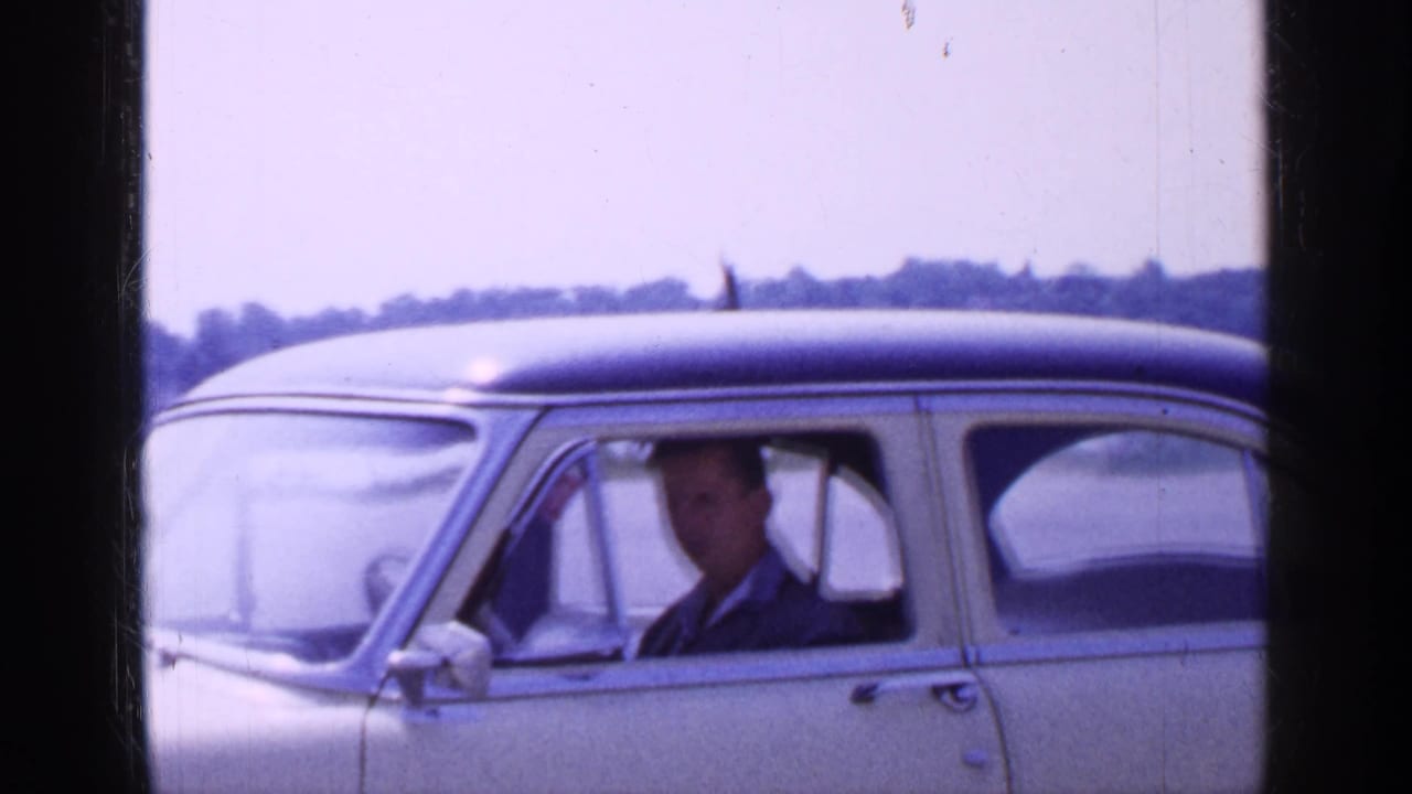 Man seated in the driver's seat of a cream sedan near a body of water.