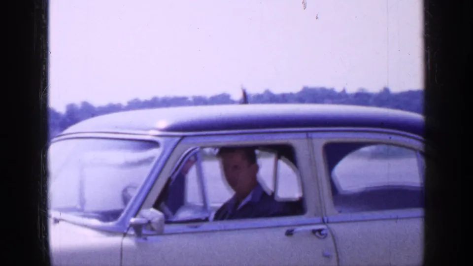 1965 Long Island frame with an adult man visible through the driver's window of a cream sedan parked near water.