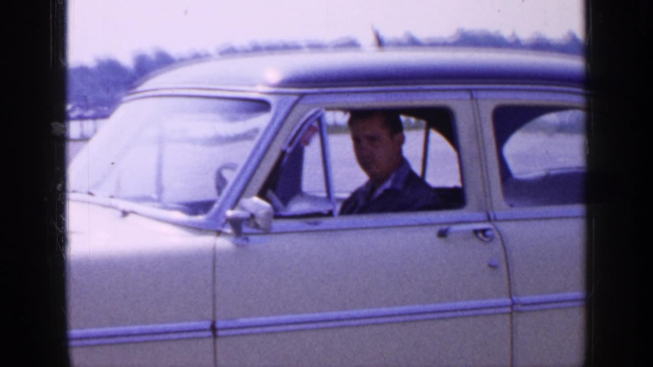 Closer 1965 Long Island home-movie view of the cream sedan, with an adult male driver looking toward the camera.
