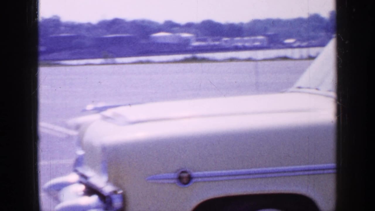 1965 Long Island frame showing the front of the cream sedan and chrome side trim near a waterfront under hazy light.