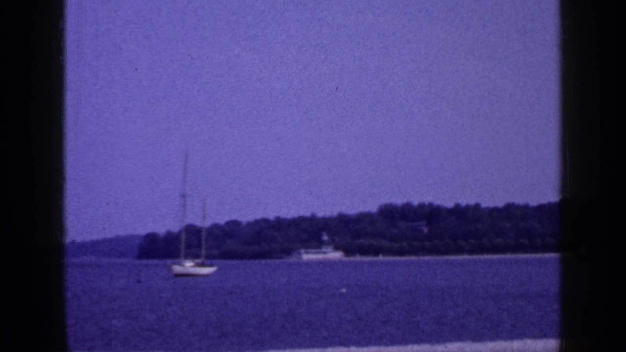 1965 Long Island waterfront view with a small sailboat offshore, wooded shoreline, and purple-shifted sky.