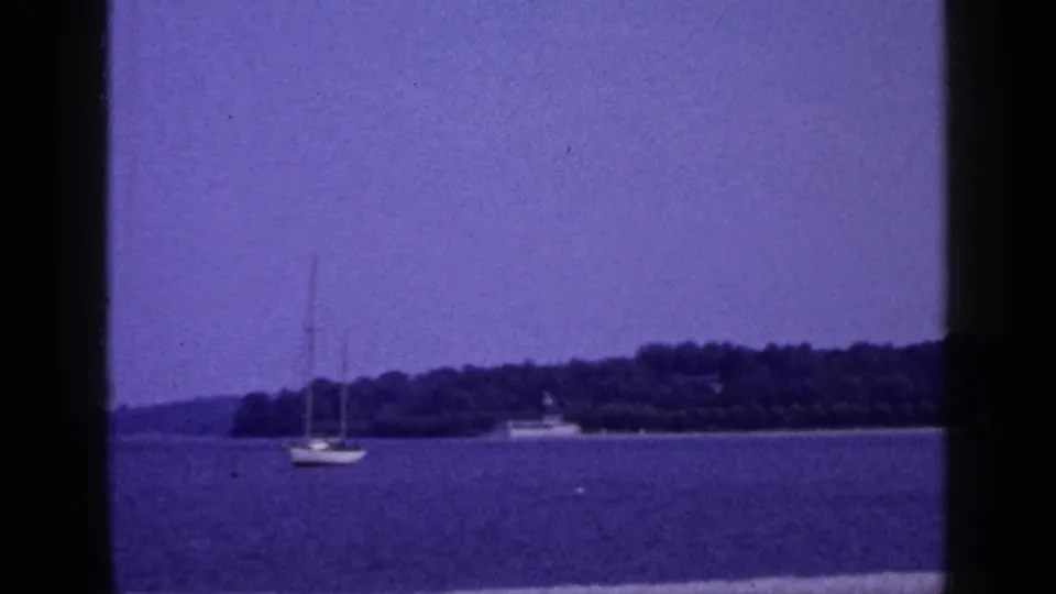 1965 Long Island waterfront view with a small sailboat offshore, wooded shoreline, and purple-shifted sky.