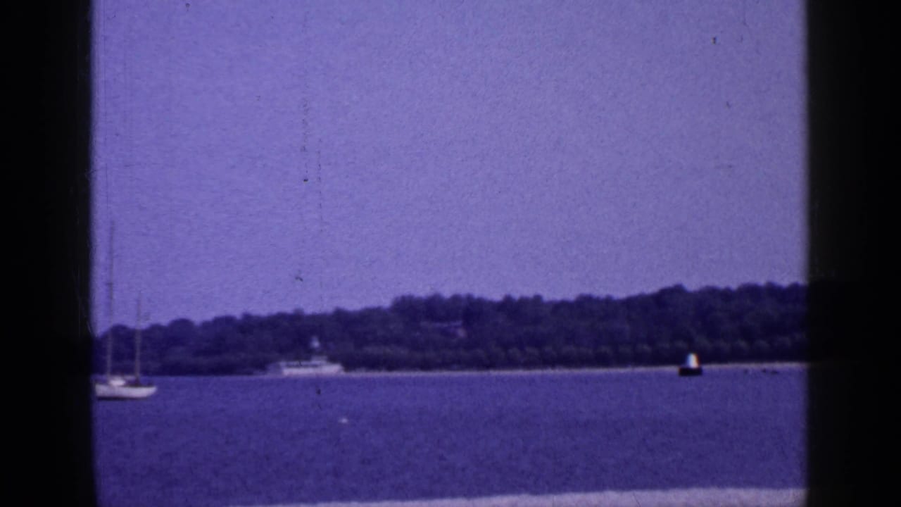 1965 Long Island lake or bay view with a sailboat at left, distant boat, buoy, and wooded shoreline.