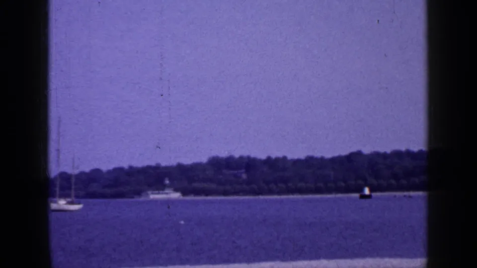 1965 Long Island lake or bay view with a sailboat at left, distant boat, buoy, and wooded shoreline.