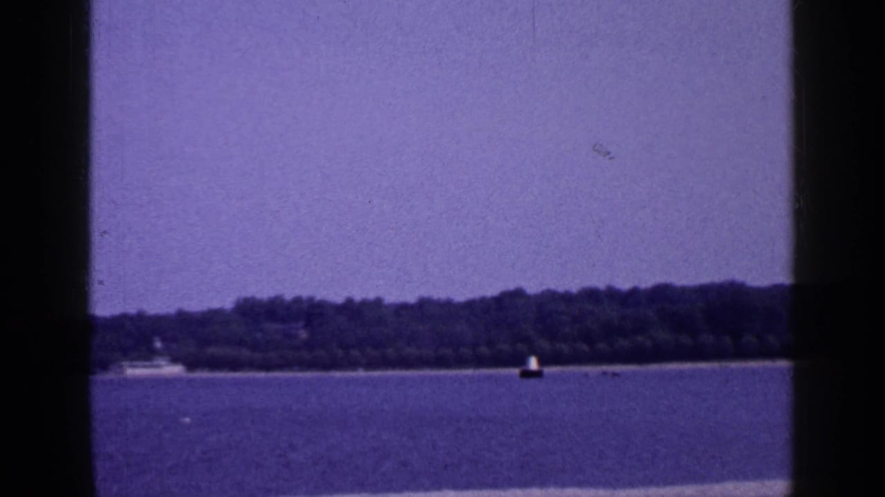 1965 Long Island home-movie frame of calm water, wooded shoreline, a small distant boat, and buoy.