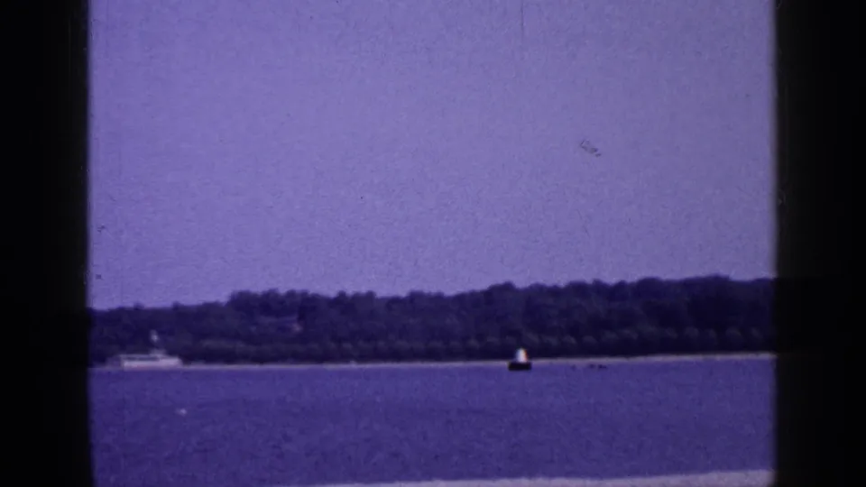 1965 Long Island home-movie frame of calm water, wooded shoreline, a small distant boat, and buoy.
