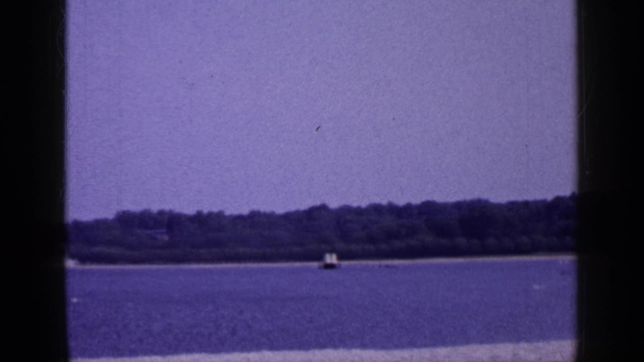 1965 Long Island 8mm frame showing calm blue water, a distant wooded shoreline, a small buoy or marker, and dark film-frame edges.