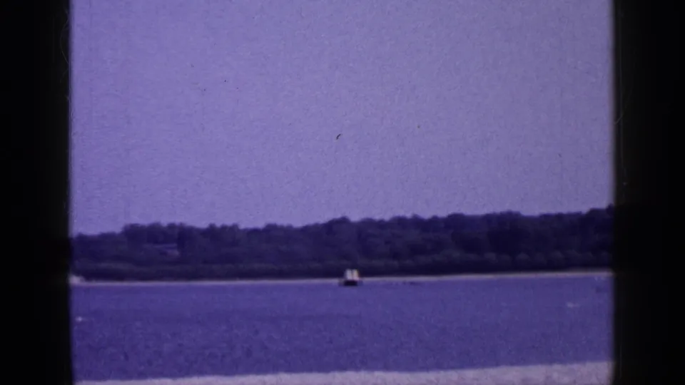 1965 Long Island 8mm frame showing calm blue water, a distant wooded shoreline, a small buoy or marker, and dark film-frame edges.