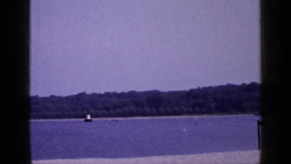 1965 Long Island beach footage frame with open water, a distant tree line, a small offshore marker, and visible 8mm frame edges.
