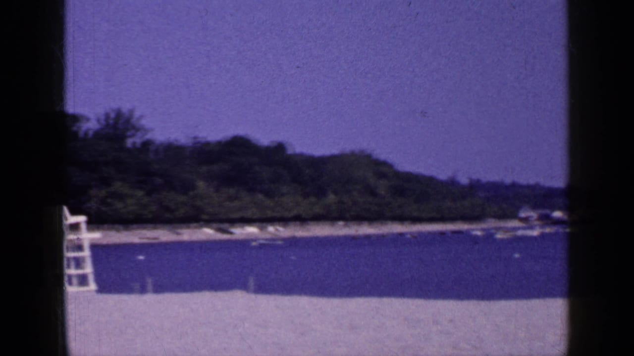 1965 Long Island shoreline frame showing pale sand, calm water, a wooded point, and a white lifeguard chair near the left edge.