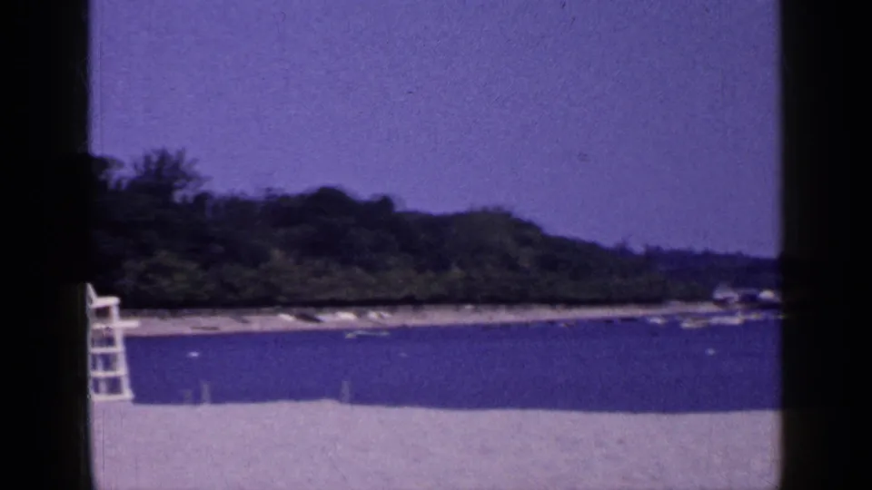 1965 Long Island shoreline frame showing pale sand, calm water, a wooded point, and a white lifeguard chair near the left edge.