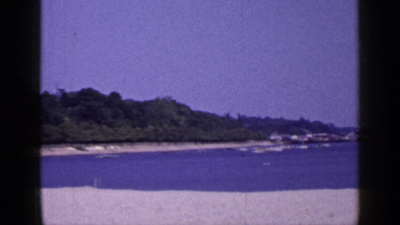 1965 Long Island 8mm frame with sand in the foreground, blue water, a low wooded shoreline, and small distant shoreline structures.