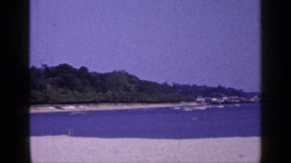 1965 Long Island 8mm frame with sand in the foreground, blue water, a low wooded shoreline, and small distant shoreline structures.