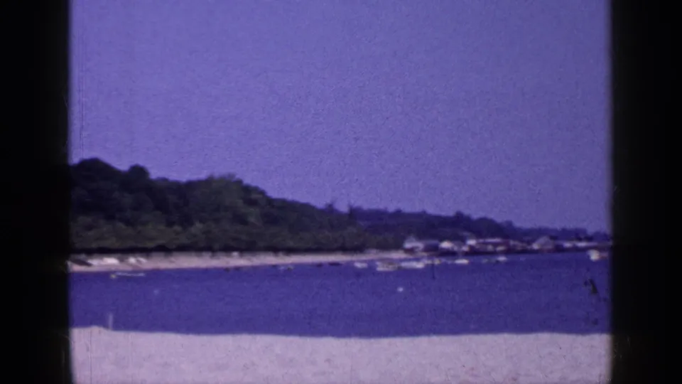 1965 Long Island coastal view with pale sand, calm blue water, anchored boats, and a tree-lined shore seen through 8mm frame edges.