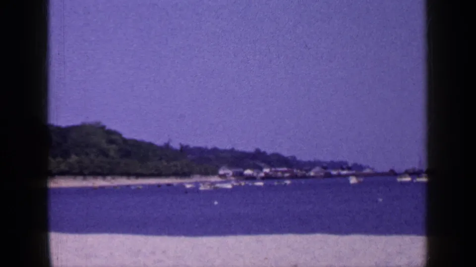 Long Island waterfront scene from 1965 showing anchored small boats, a sandy foreground, distant trees, and low shoreline structures.