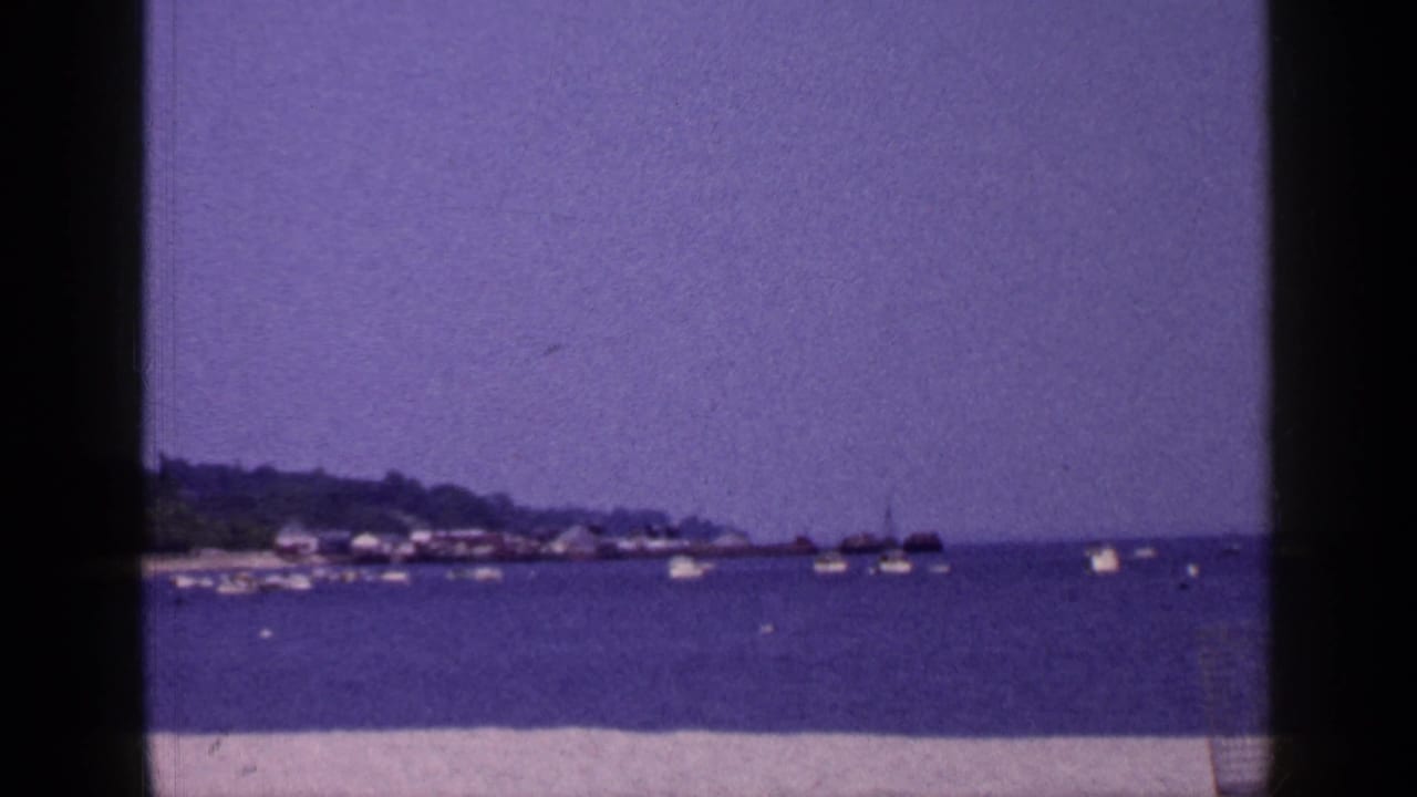 Long Island coastal home-movie frame from 1965 with sand in front, boats offshore, and a low tree-lined shoreline under open sky.