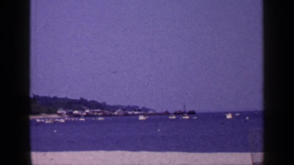 Long Island coastal home-movie frame from 1965 with sand in front, boats offshore, and a low tree-lined shoreline under open sky.