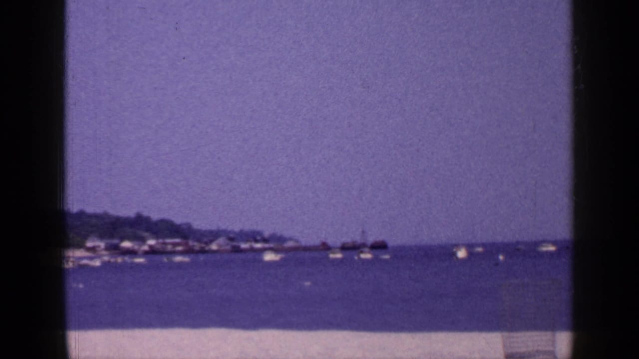 1965 Long Island 8mm view of a calm coastal harbor with boats, hazy sky, distant shoreline, and pale sand in front.