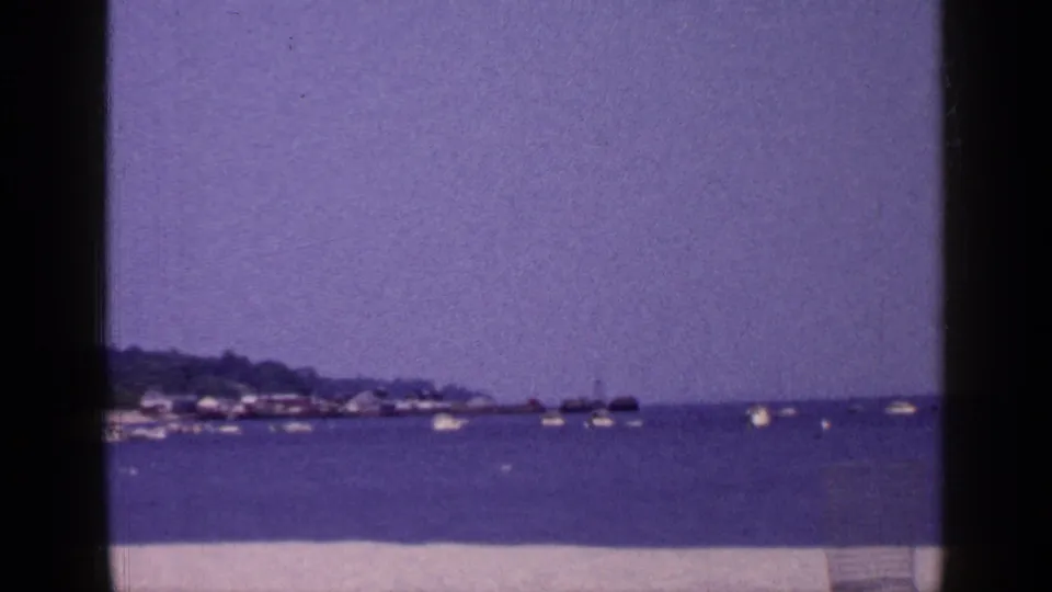 1965 Long Island 8mm view of a calm coastal harbor with boats, hazy sky, distant shoreline, and pale sand in front.