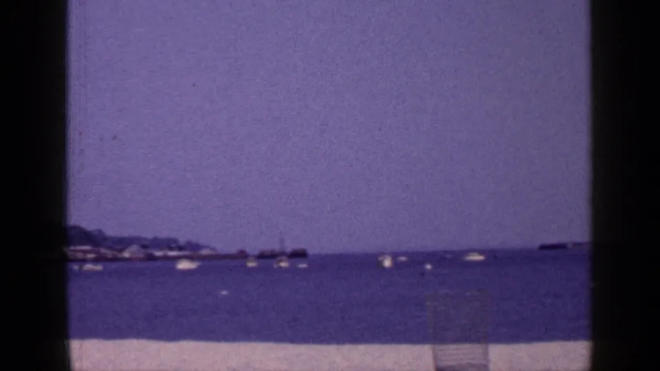 1965 Long Island coastal harbor scene with scattered boats on still water and a low shoreline under a purple-tinted sky.