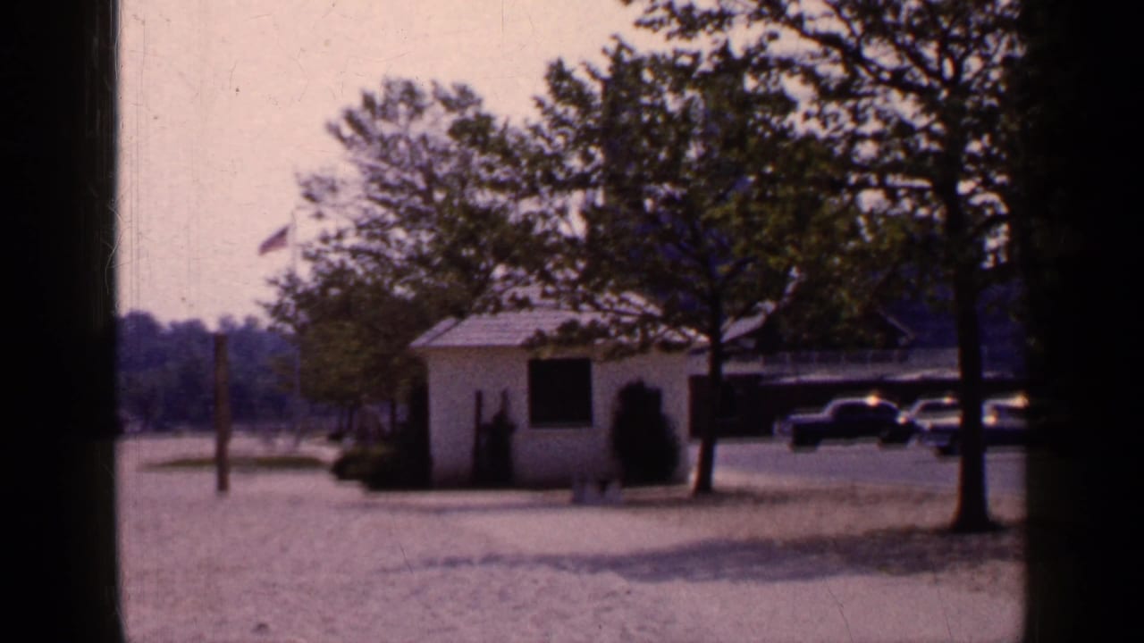 1965 Long Island beachside area with a small white hut, trees, sandy ground, and parked cars near the waterfront.