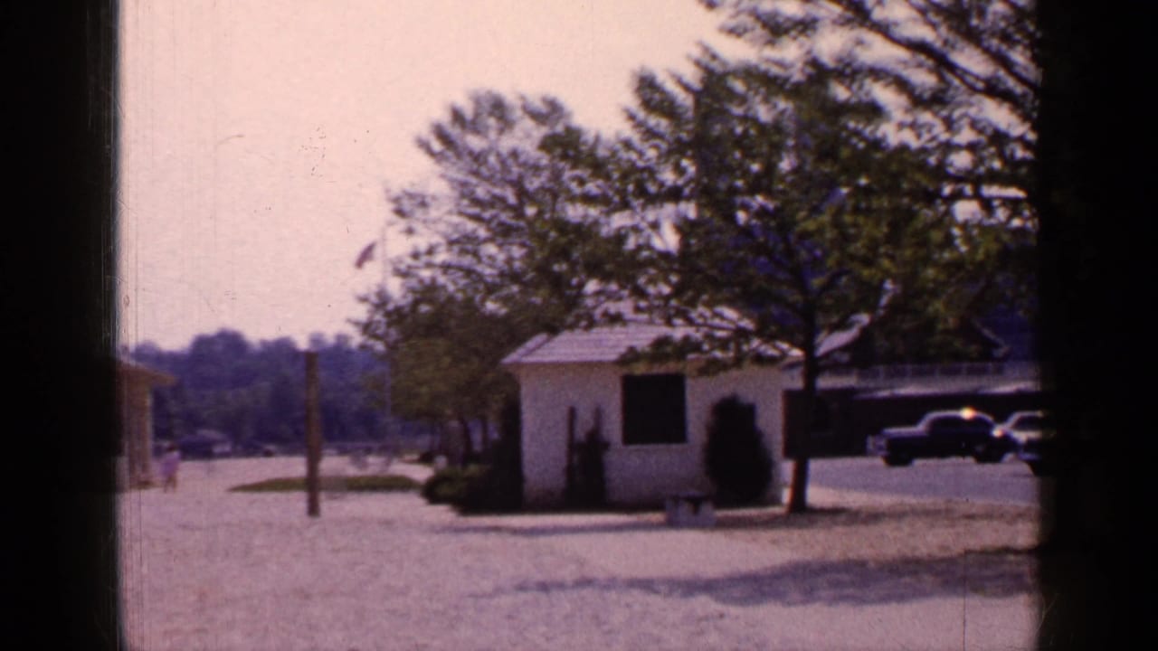 1965 Long Island park view with a white shed, trees, gravel foreground, partial yellow gazebo, parked cars, and soft 8mm film color.