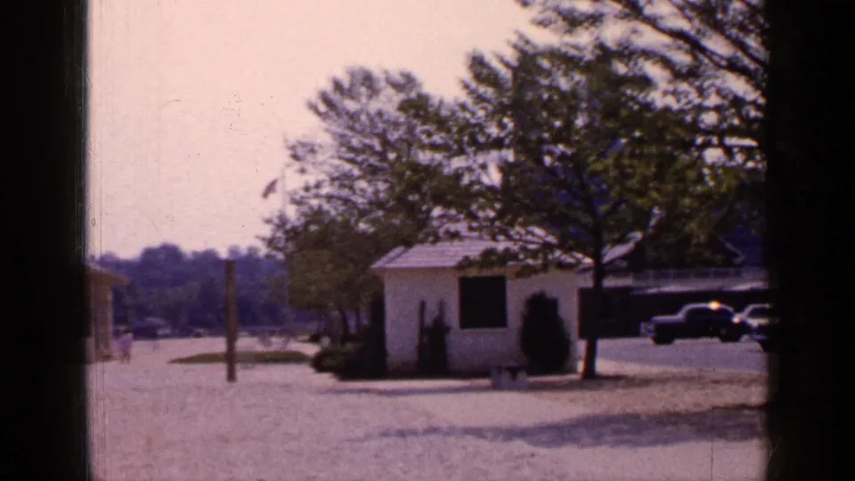 1965 Long Island park view with a white shed, trees, gravel foreground, partial yellow gazebo, parked cars, and soft 8mm film color.