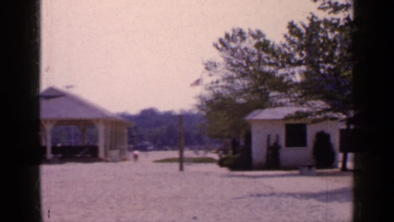 Yellow gazebo beside a small white shed, with trees, gravel, parked cars, and a flag in the background.