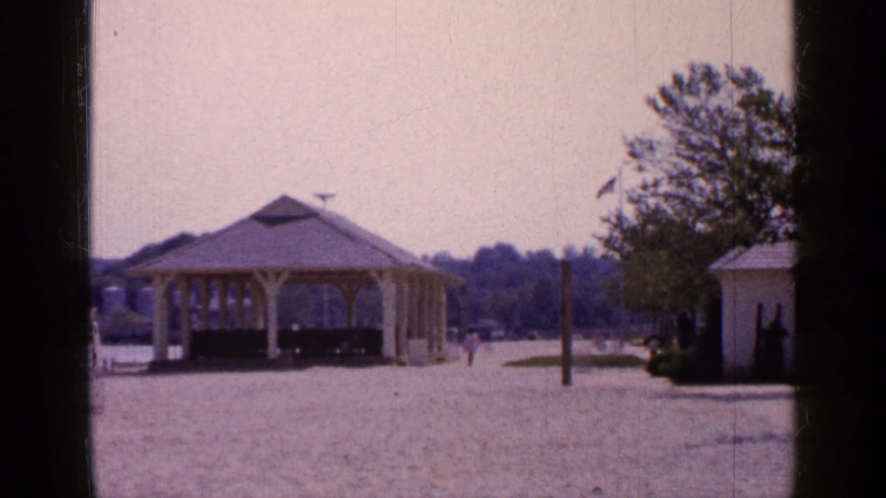 1965 Long Island park view with the yellow gazebo centered, a white shed to the side, a flagpole, trees, and small distant pedestrians.