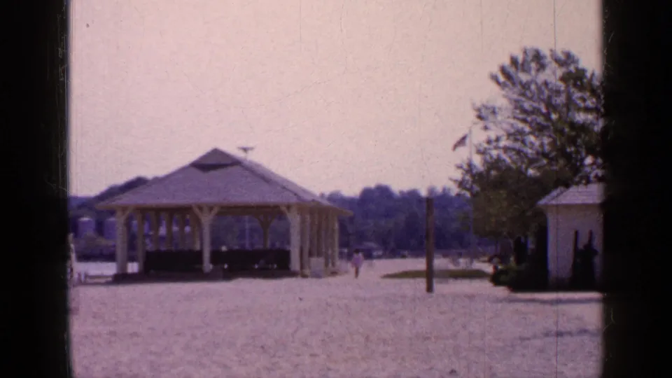 1965 Long Island park view with the yellow gazebo centered, a white shed to the side, a flagpole, trees, and small distant pedestrians.