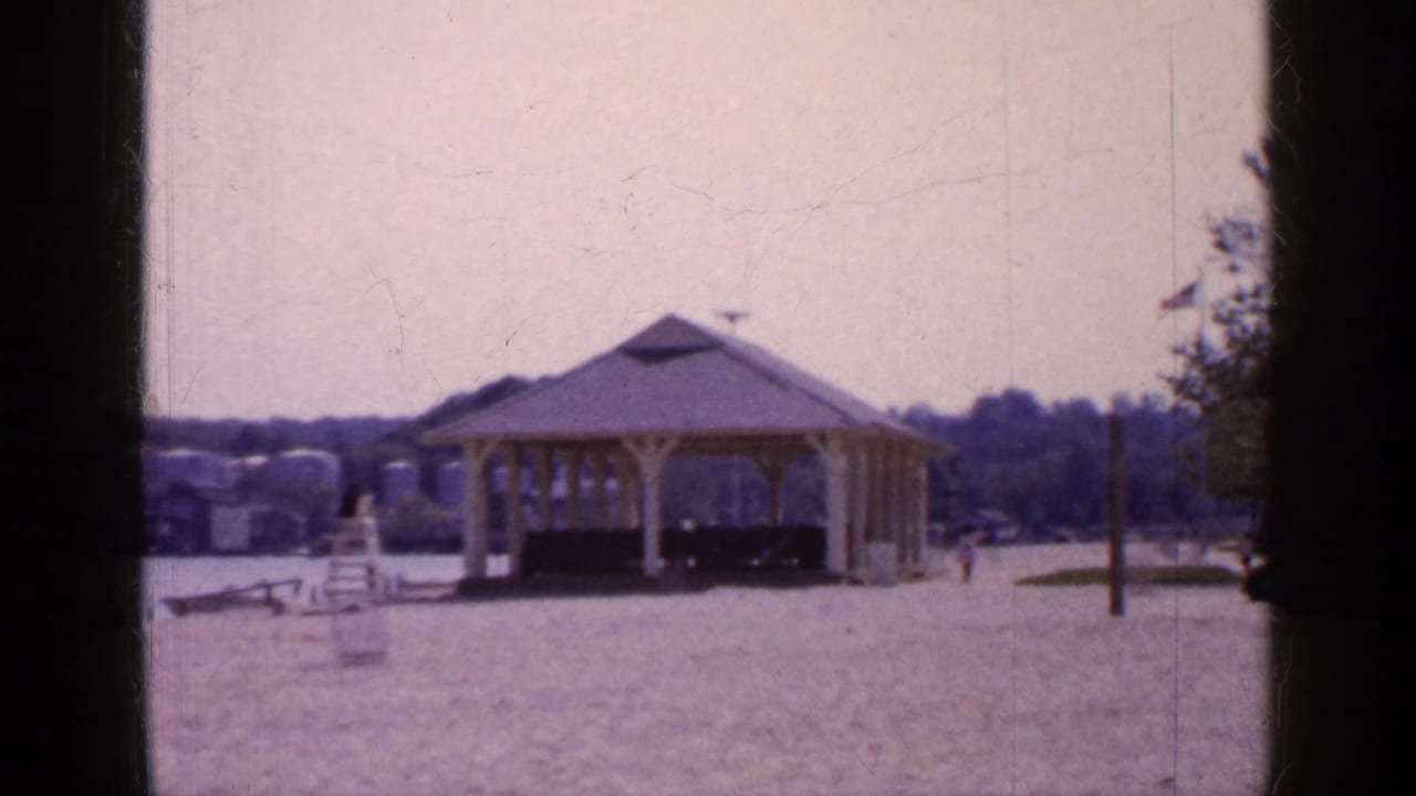 1965 Long Island park frame with the yellow wooden gazebo, open gravel foreground, flagpole, trees, distant pedestrians, and water in the background.