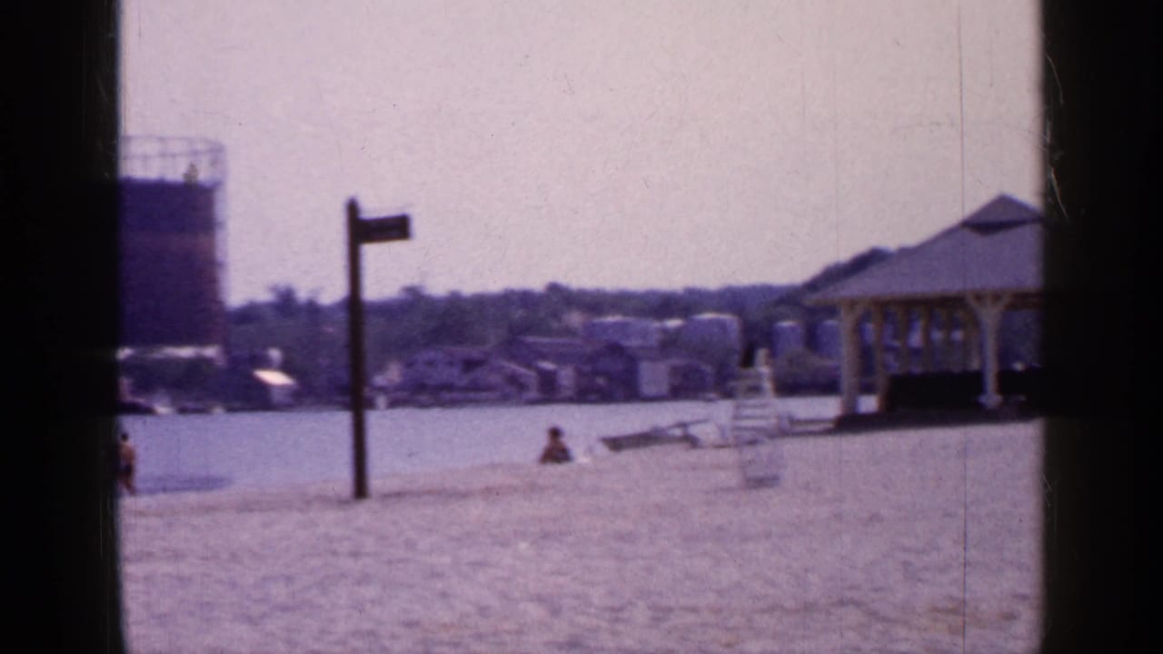 Sandy waterfront with a small shelter, distant houses, a large tank, and dark film gate edges.