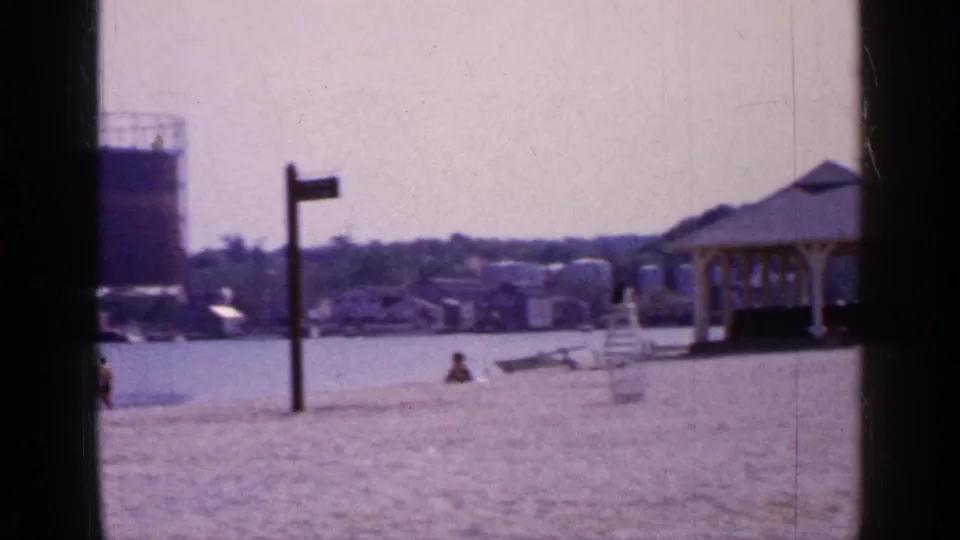 1965 Long Island waterfront view with sandy beach, small shelter, distant shoreline houses, utility poles, and a large tank at left.
