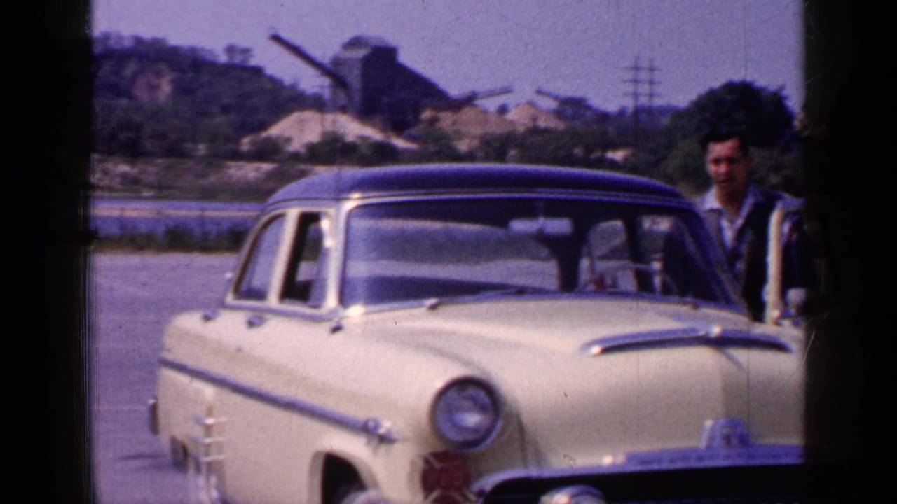 Yellow mid-century sedan seen from the front with an adult man partly visible beside the driver side at the waterfront location.