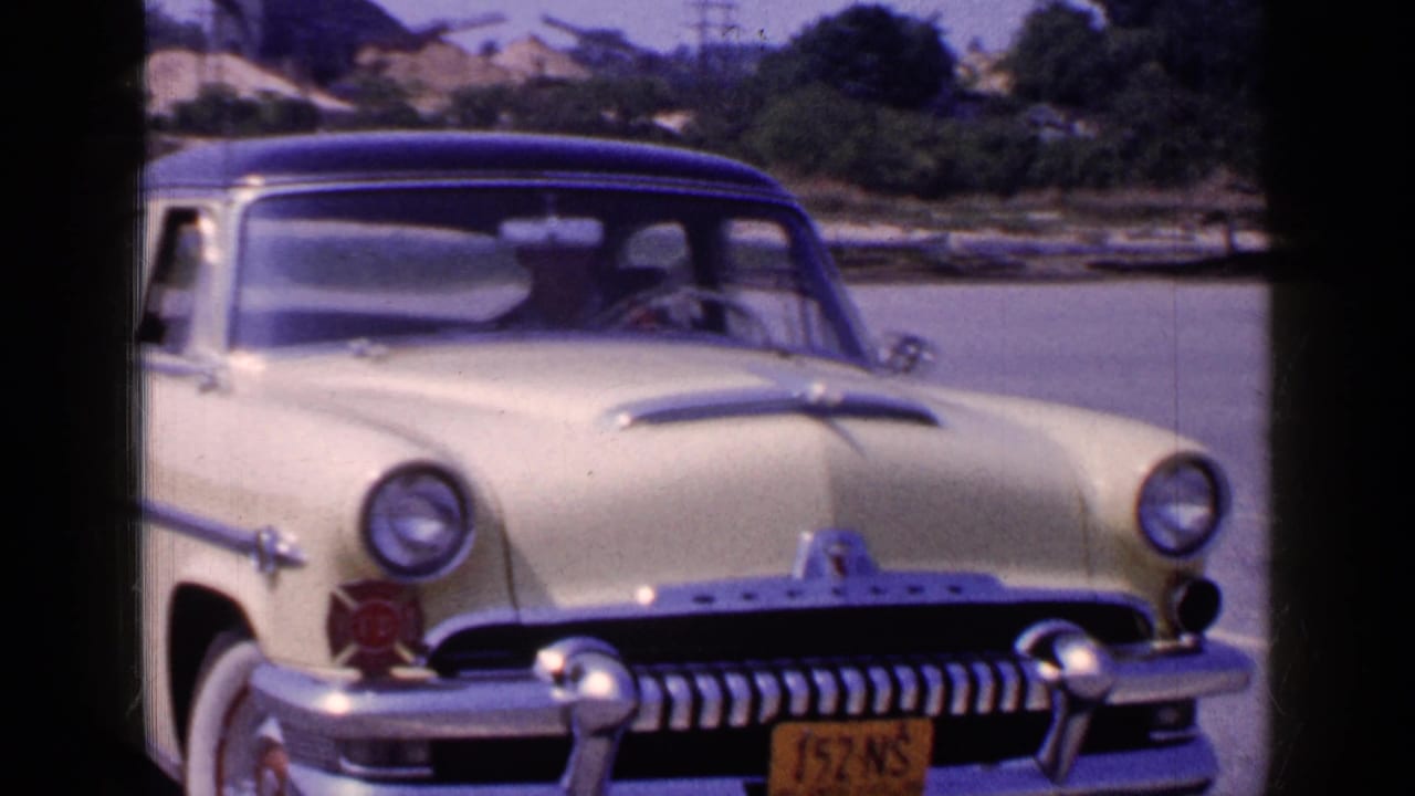 Cream classic sedan with dark roof parked near water, driver visible through the windshield.