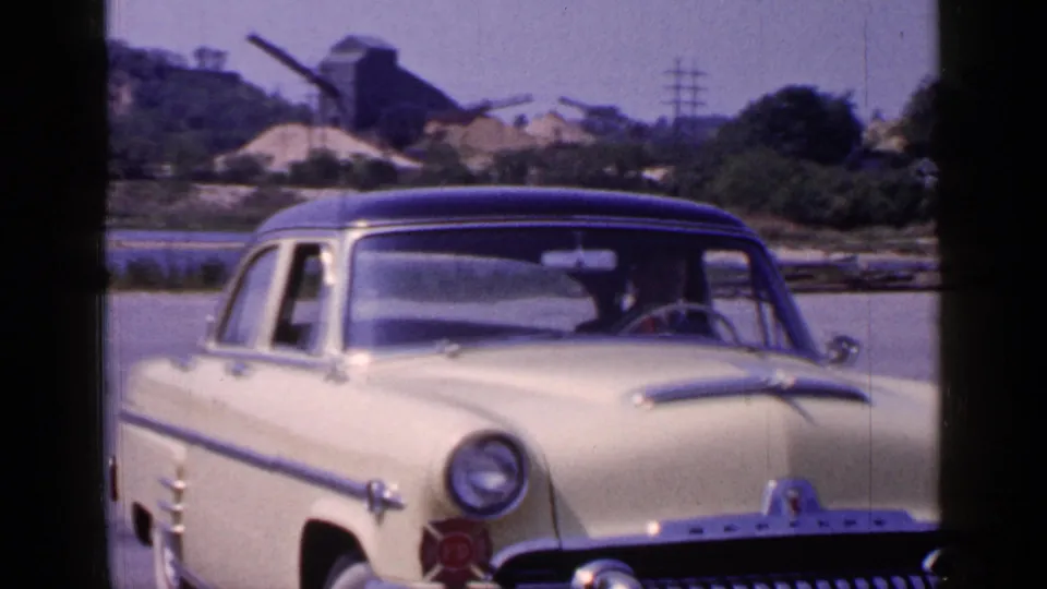 1965 Long Island frame with a cream mid-century sedan, dark roof, chrome bumper, driver, waterfront, trees, and distant industrial structures.
