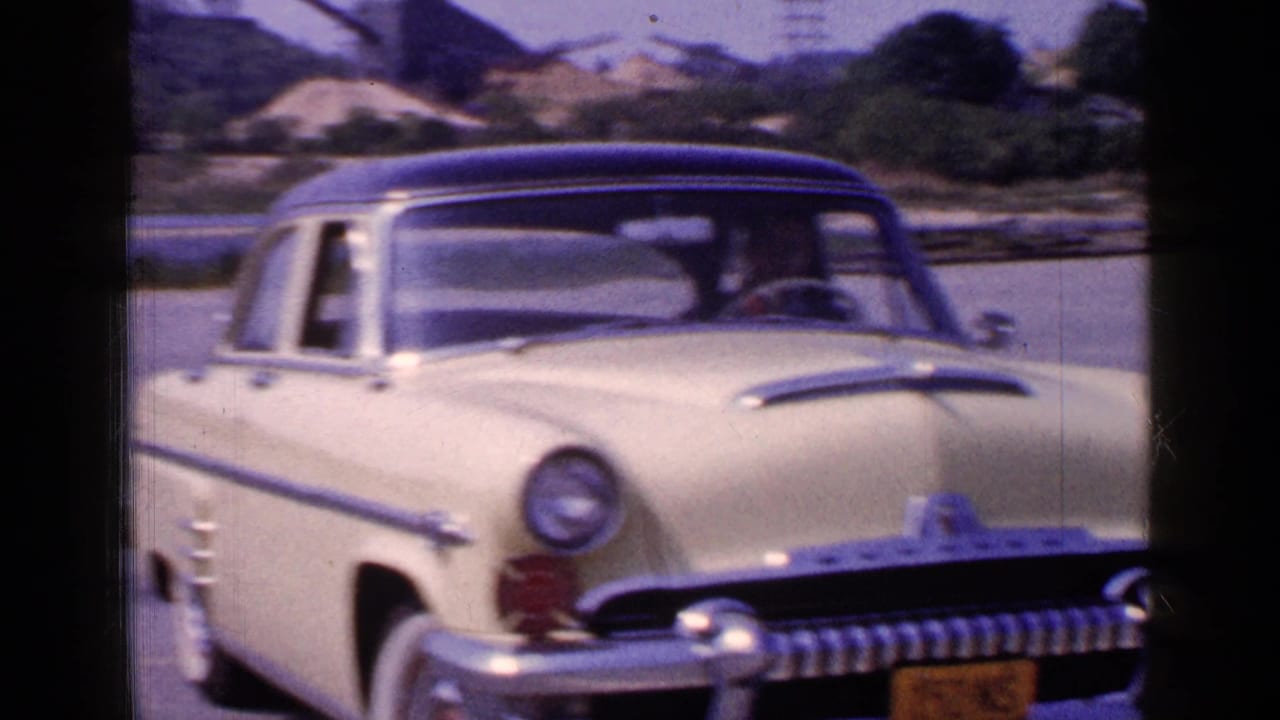1965 Long Island home-movie view of the cream sedan from a closer angle, with chrome trim, yellow plate, driver, water, and shoreline behind it.