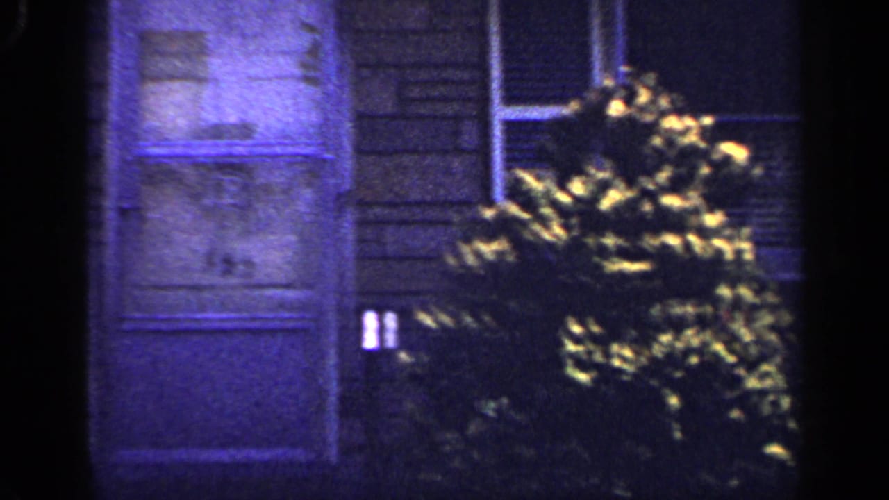 8mm home-movie frame showing a Long Island suburban doorway marked 152, adjacent brick wall, window edge, and garden shrubs.