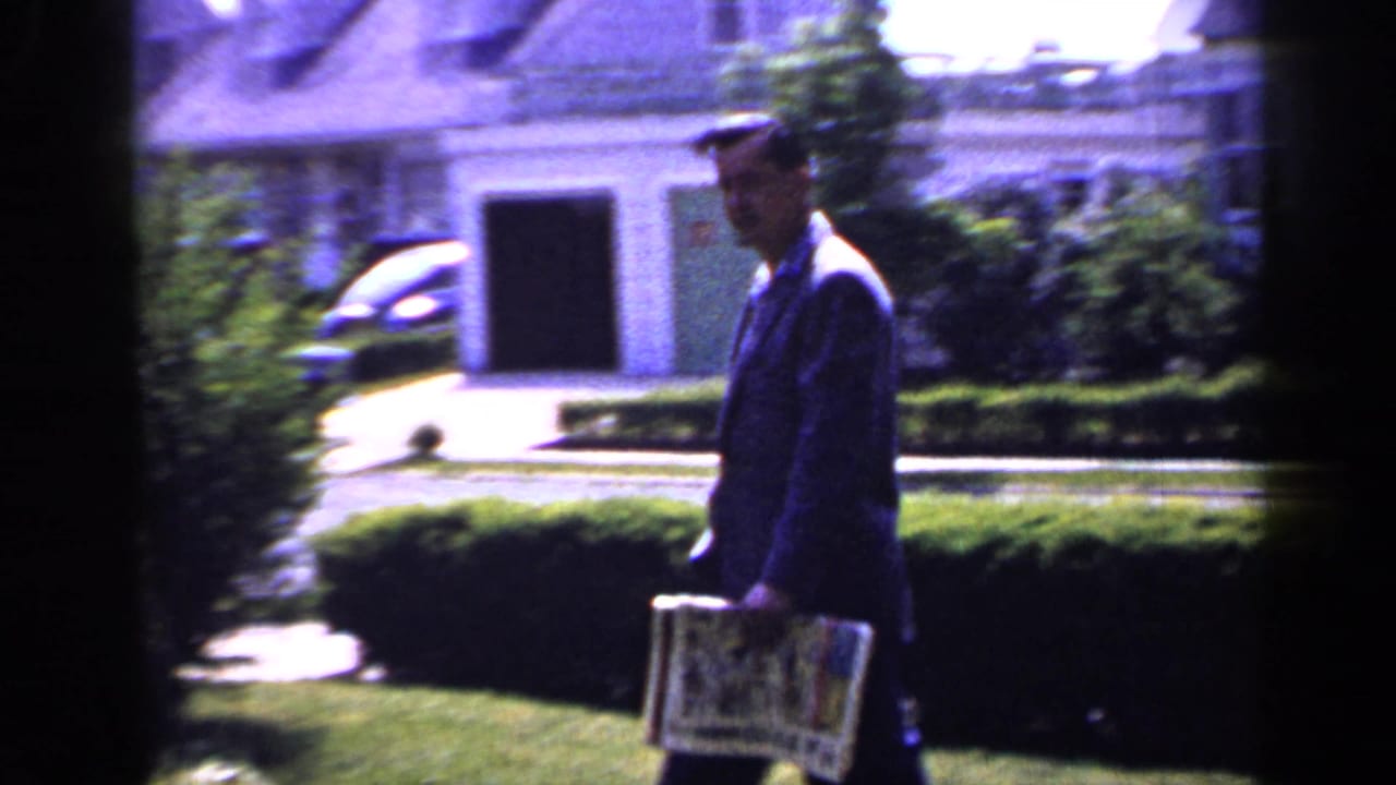 In 1965 Long Island home-movie footage, a suited man carries a newspaper past lawn, hedges, and a house exterior.