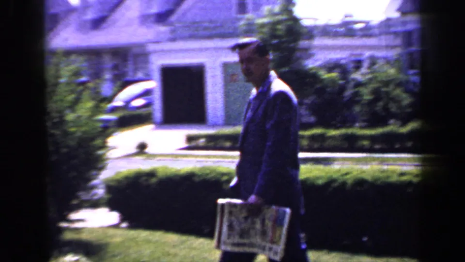 In 1965 Long Island home-movie footage, a suited man carries a newspaper past lawn, hedges, and a house exterior.