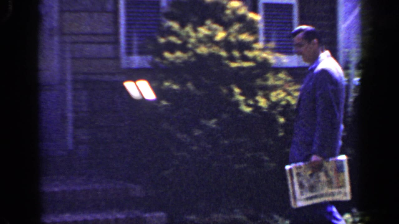 The suited man continues along a Long Island residential exterior, holding a newspaper near bushes and shuttered windows.