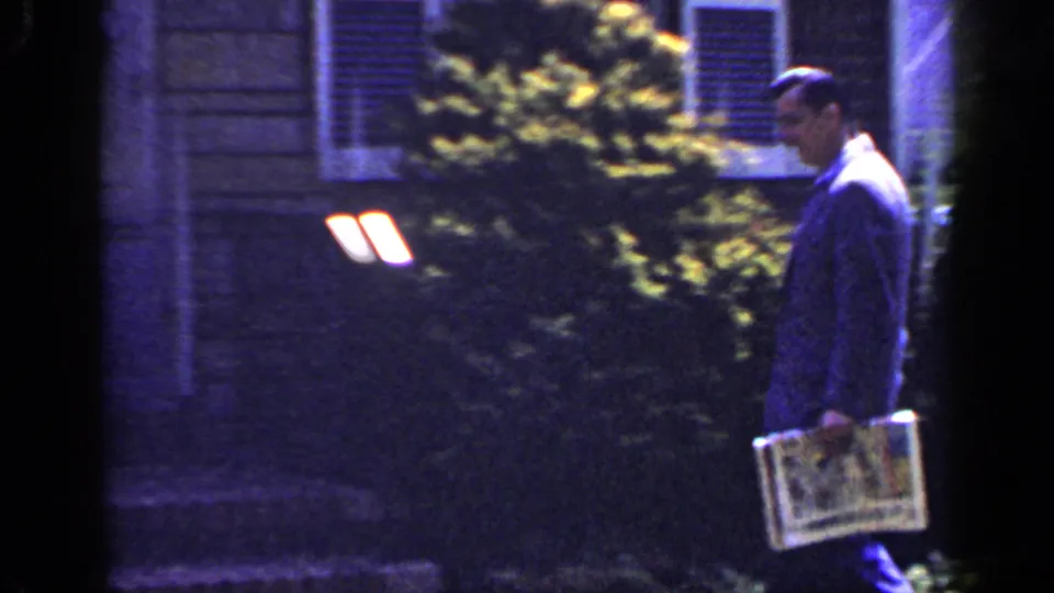 The suited man continues along a Long Island residential exterior, holding a newspaper near bushes and shuttered windows.