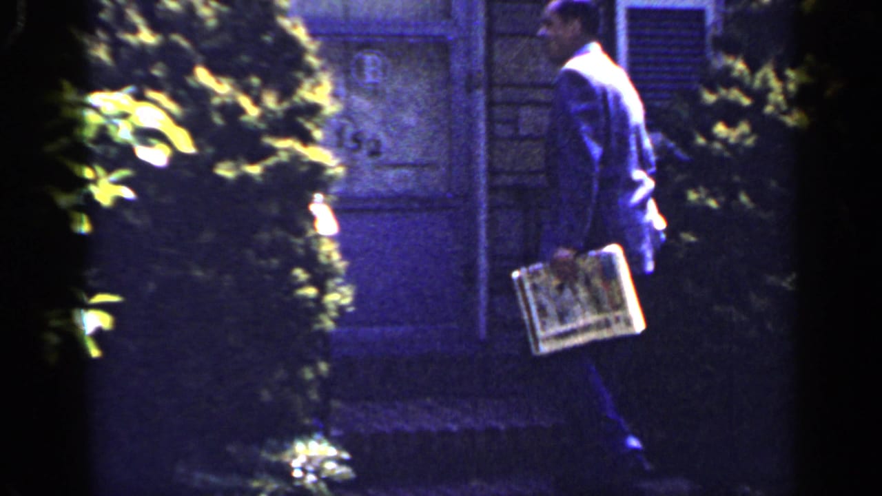 The home-movie frame shows the man from behind, carrying a newspaper along a residential path in Long Island.