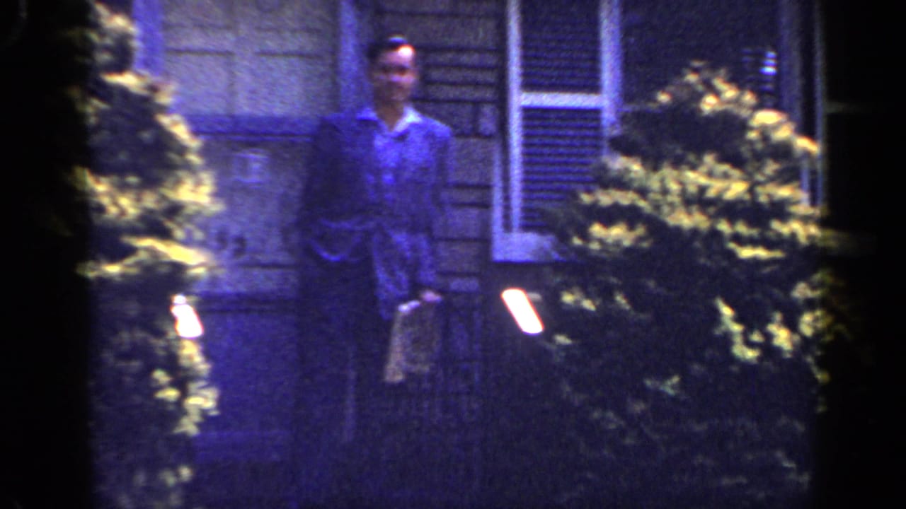 Man in a suit stands near a house entrance with shrubs in the foreground.