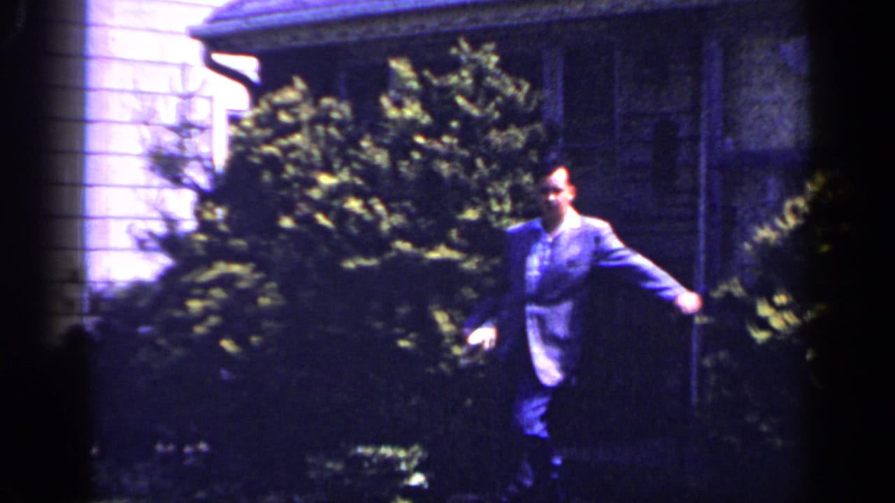 Adult man in a suit moves across the yard outside the Long Island house, with shrubs and porch area behind him.