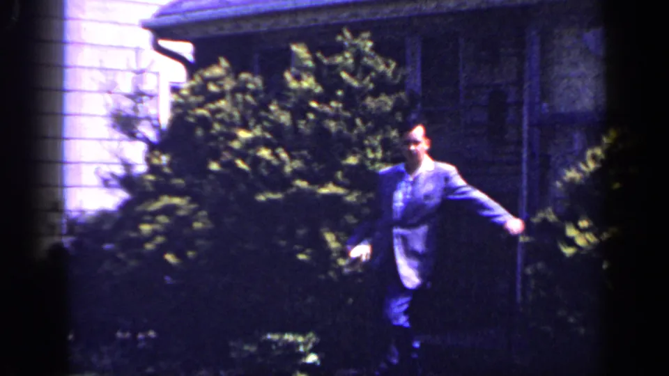 Adult man in a suit moves across the yard outside the Long Island house, with shrubs and porch area behind him.