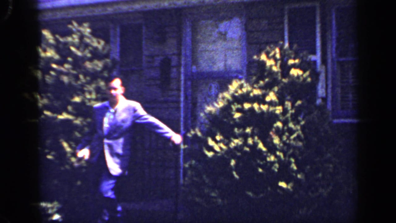 In the 1965 Long Island home-movie frame, the suited man continues across the yard with house windows and plantings behind him.